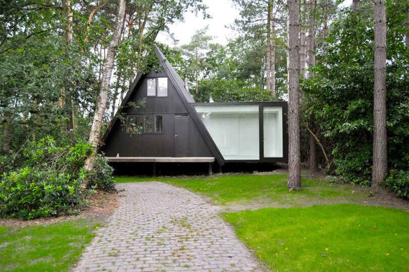 front exterior of black a-frame cabin in woods with white extension