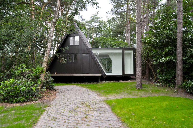 front exterior of black a-frame cabin in woods with white extension