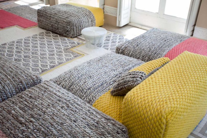 open room with windows filled with low pouf seating in pink, yellow, and greys along with rugs