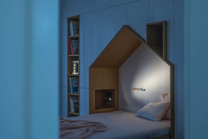 children's bedroom with house-shaped bed embedded into wall of white built-in storage units at night