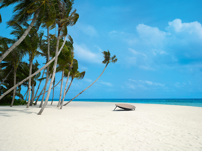 um salão em forma de orbe à distância em uma praia com areia, palmeiras e céu azul