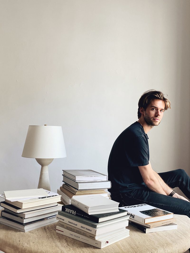 light-skinned man wearing black and sitting on table with stacks of books and lamp