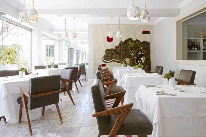 interior dining space with white tablecloth covered tables, armchairs and light walls and floor