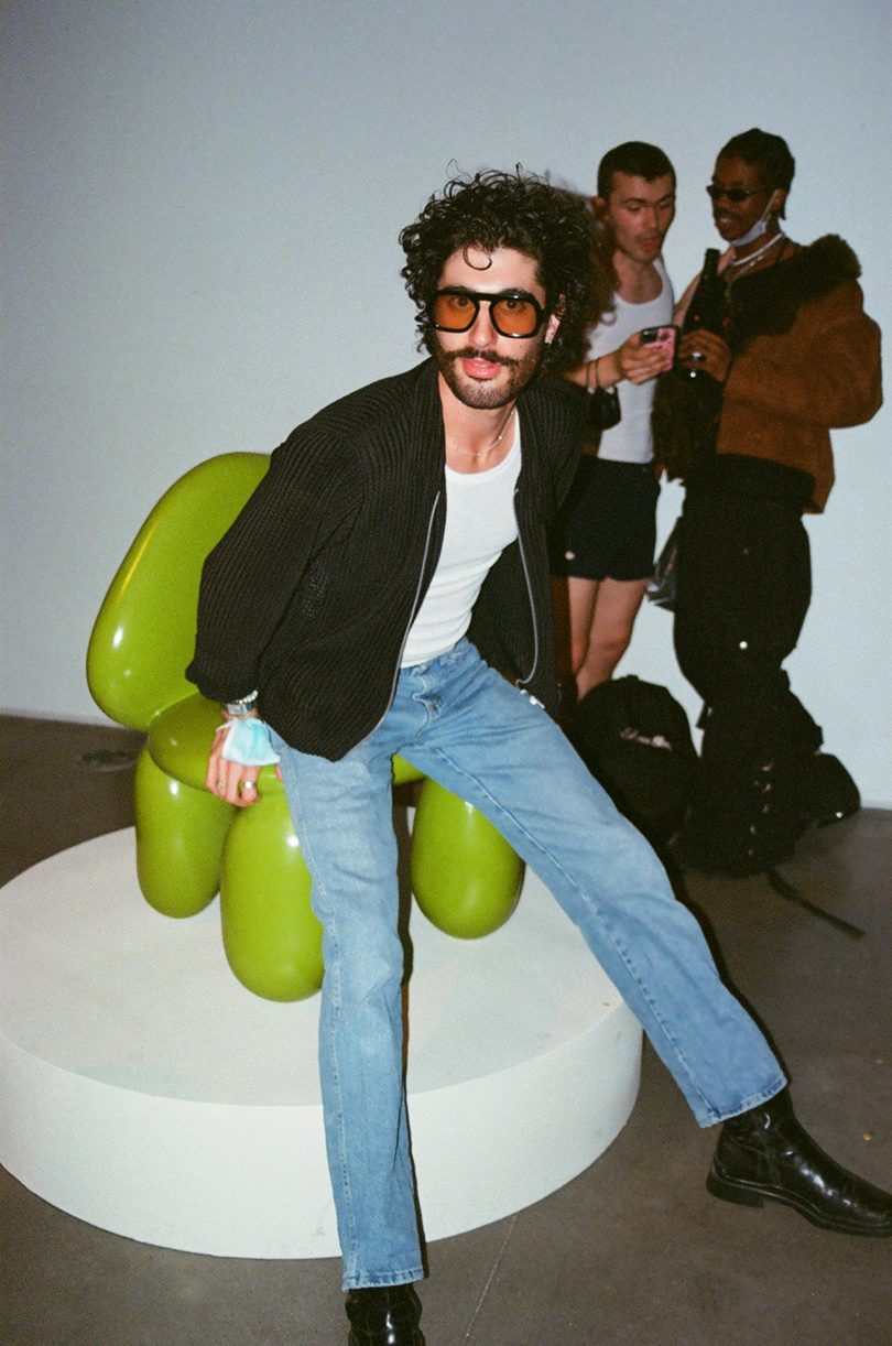 light-skinned man with dark hair wearing a leather jacket, jeans, and sunglasses