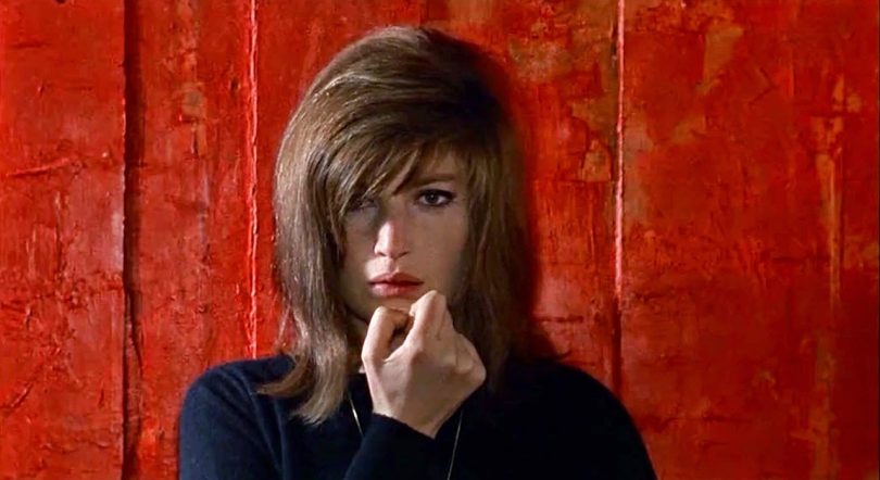 film still of slight skinned woman with medium length brown hair and black shirt in front of red wall