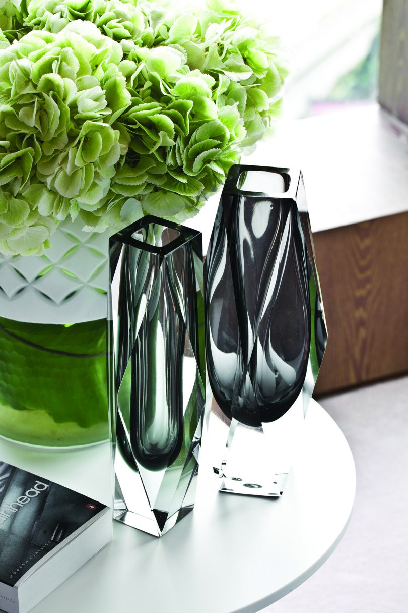 two empty glass vases next to one another with flowers behind them