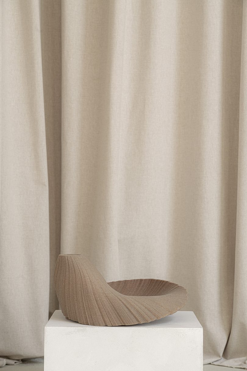 light brown abstract vessel sitting on white pedestal in front of light fabric