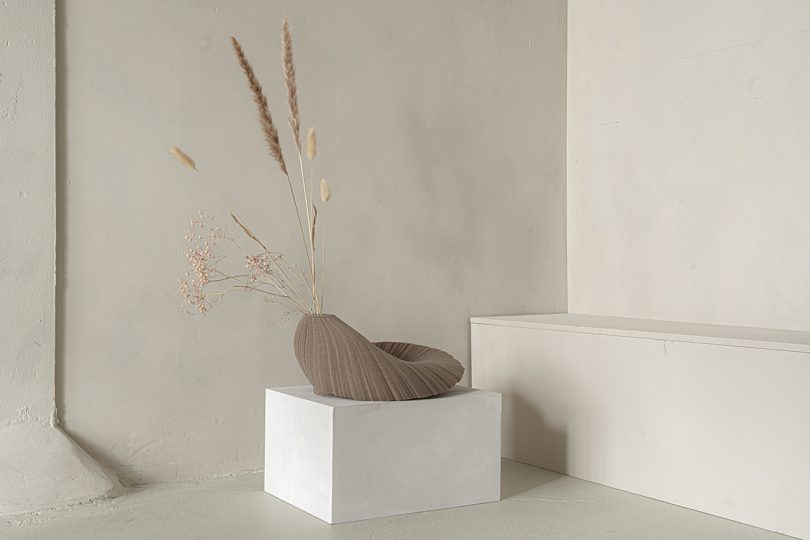 light brown abstract vessel sitting on white pedestal in front of light wall
