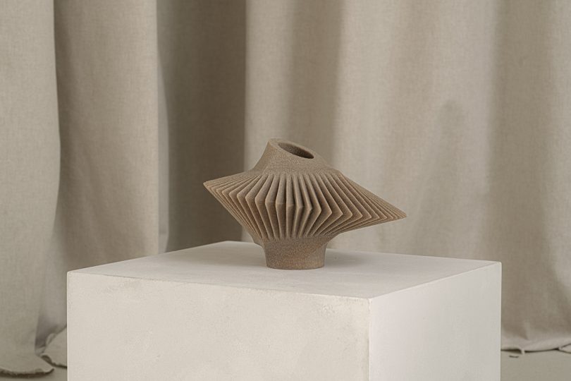 light brown abstract vessel sitting on white pedestal in front of light fabric