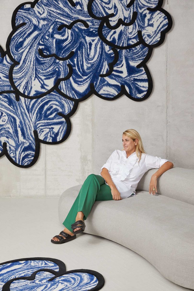 light skinned woman with light hair sitting on a white sofa below a nebulous shaped blue floor rug hung on white wall