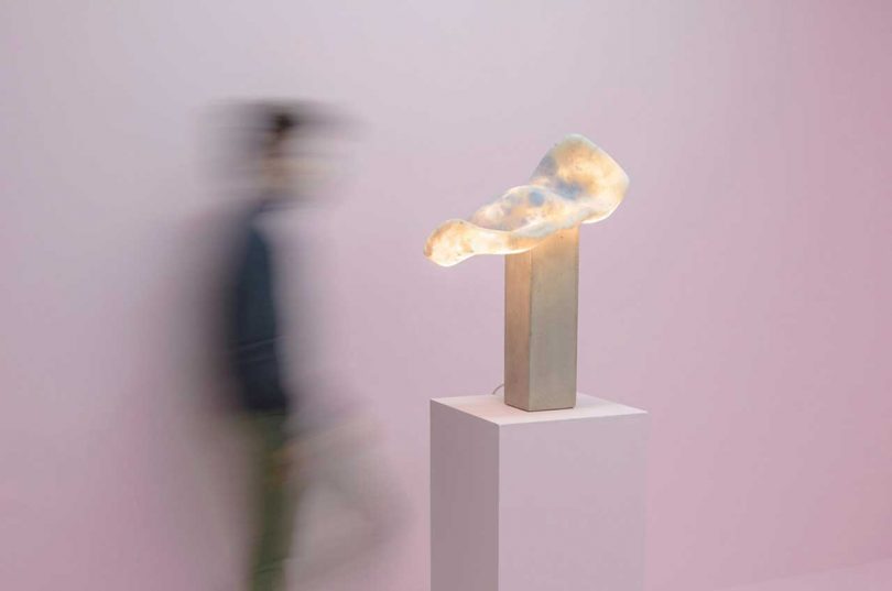 abstract sculptural table lamp on pedestal in front of light pink wall with person walking by in the background
