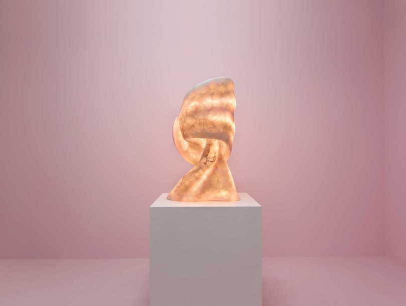 lit abstract sculptural table lamp on pedestal in front of light pink wall