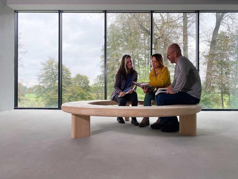 three people sitting together on circular wood bench in front of windows