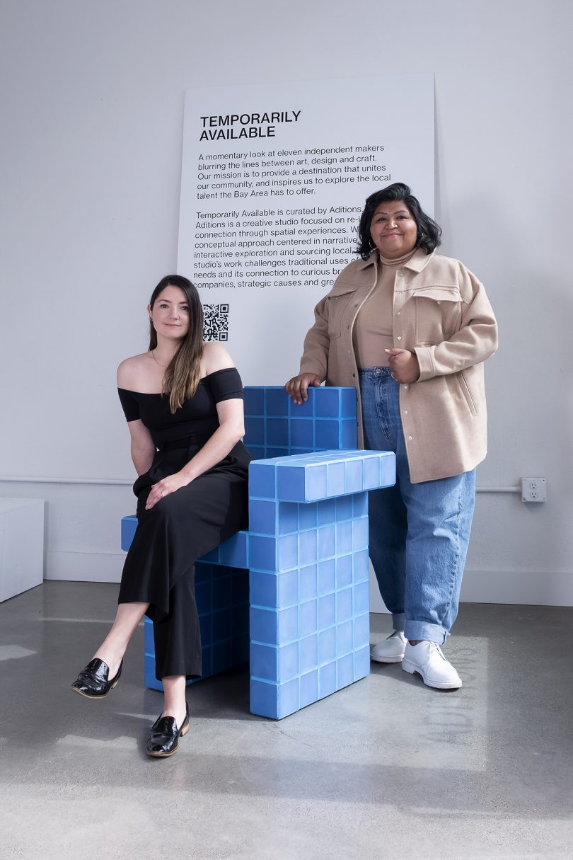 two women posing with a blue chair in a white gallery space