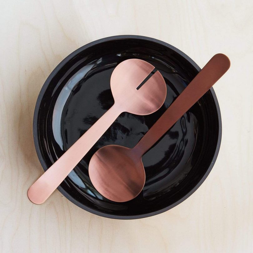 Aaron Probyn Zofia Salad Servers on top of a black bowl on a light wooden surface