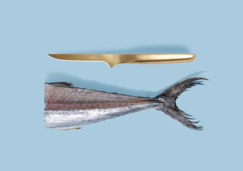 Hast knives bold gold boning knife next to half of a fish body on a blue background