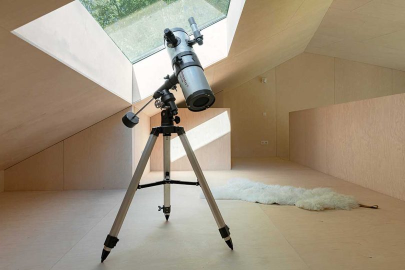 light wood paneled interior space with slanted roof with telescope pointed towards roof windows
