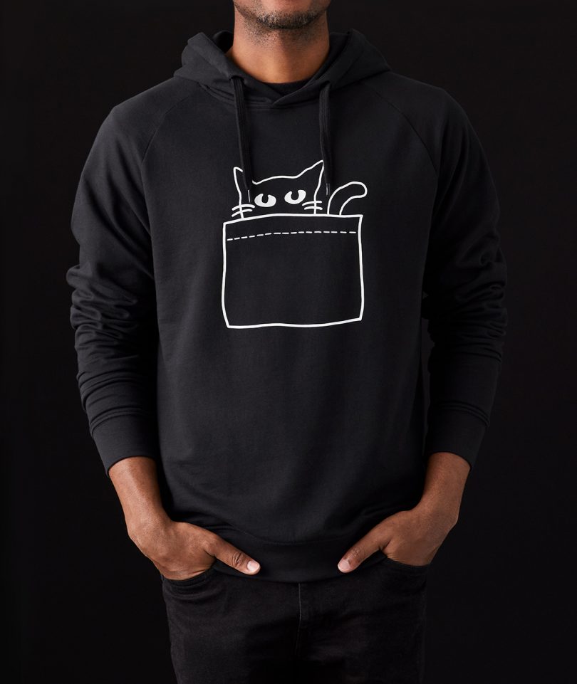 person with light-brown skin wearing black hoodie sweatshirt with cat peeking out of pocket illustration on black background