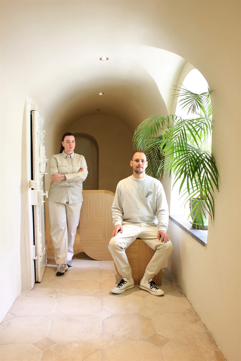 two light-skinned people wearing neutral clothing in a hallway with marquetry sculptures and potted palm