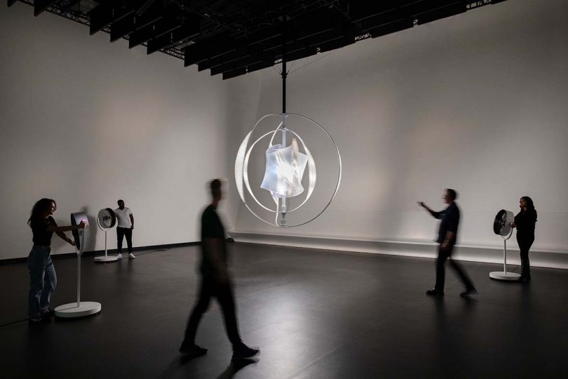The "We Harvest Wind" sculpture suspended by a single pole within a room, surrounded by three fans being pointed toward the sculpture to make it move.