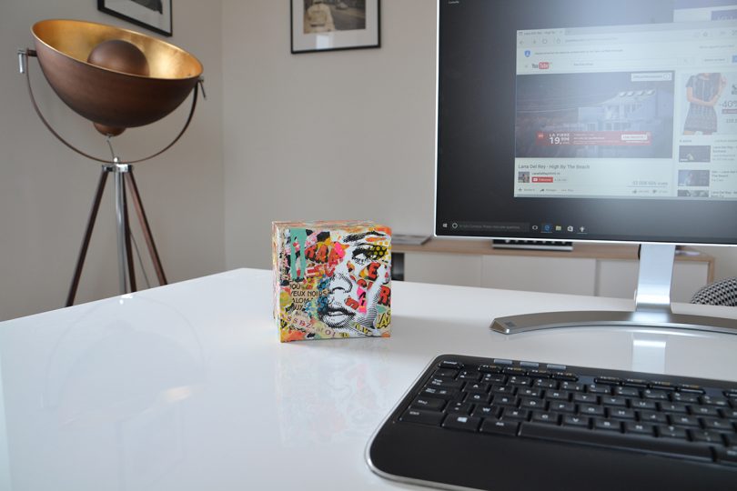 Angled view of cube shaped PC with colorful exterior case on desk with monitor and mouse, floor lamp in background.