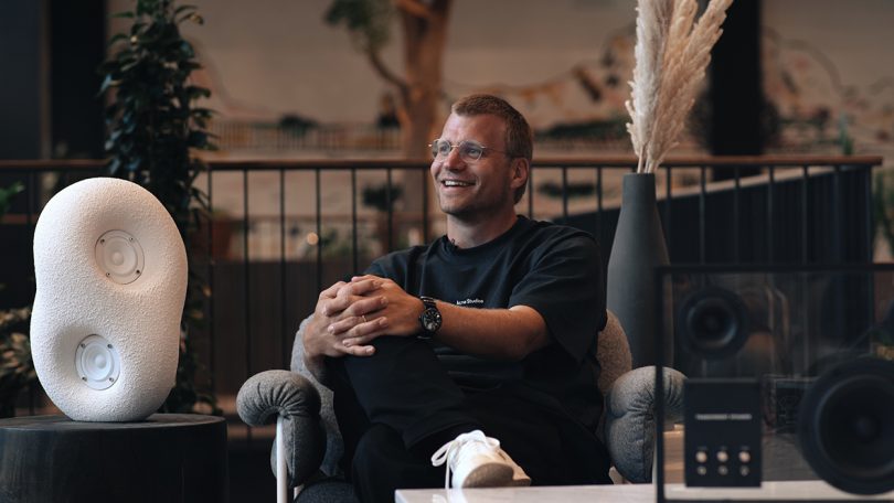 light-skinned man wearing glasses and black clothing sitting in an armchair