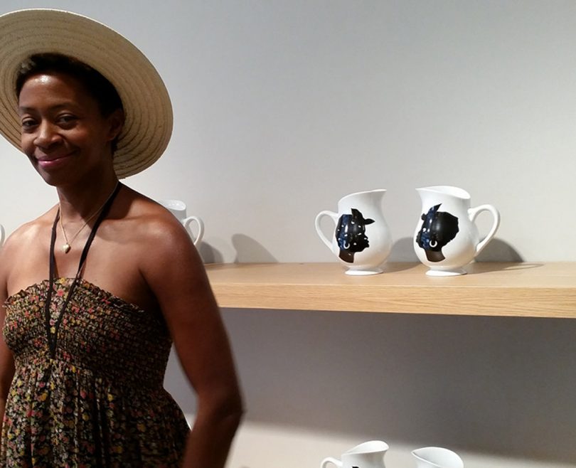 dark-skinned woman wearing dark halter dress and brimmed hat standing in front of two white and black vases