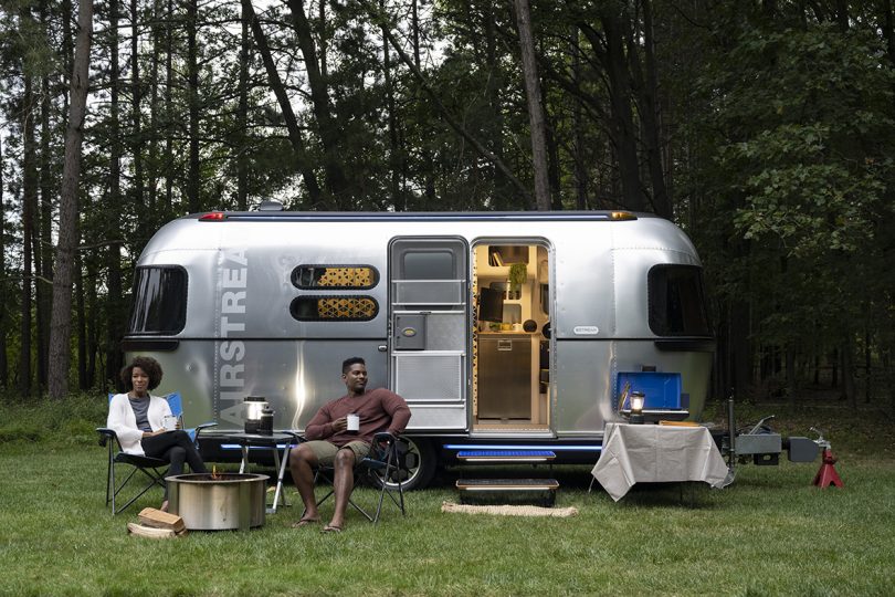 Black woman and man relaxing in chairs in front of Airstream eStream trailer parked in woods.