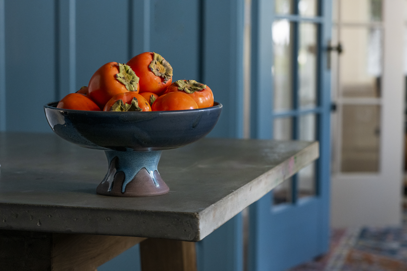 blue ceramic pedestal bowl with persimmons on a farm table