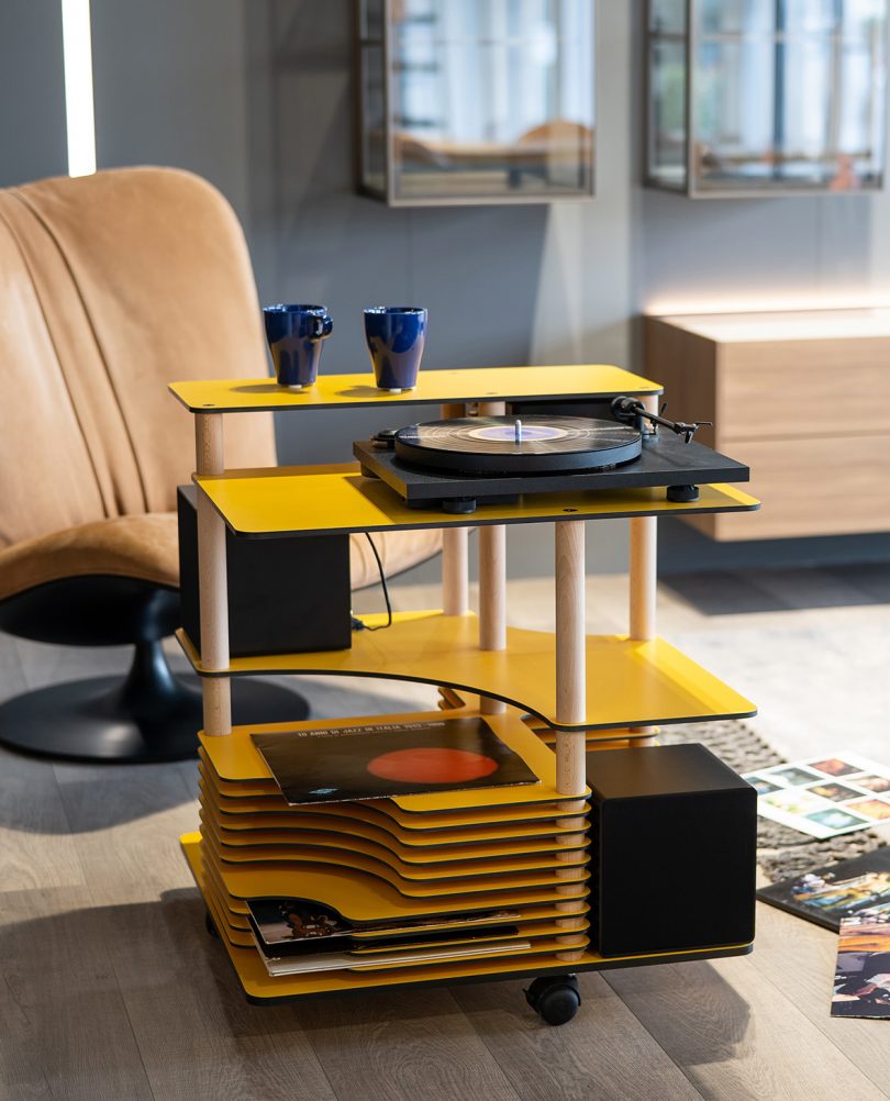 View of multitiered record stand cart in yellow with lounge chair behind it.