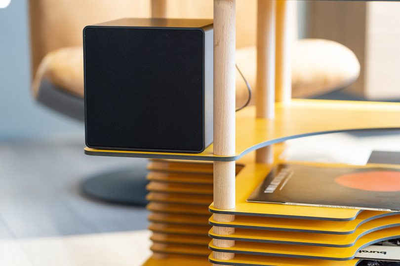 Detail of black cube speaker sitting on top of platform stand as part of yellow laminate shelf.