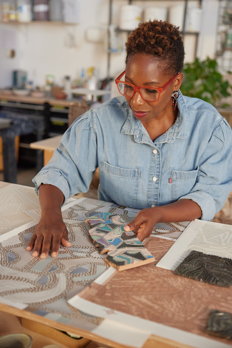 dark-skinned woman wearing red glasses and chambray shirt examining patterned wallpaper on table