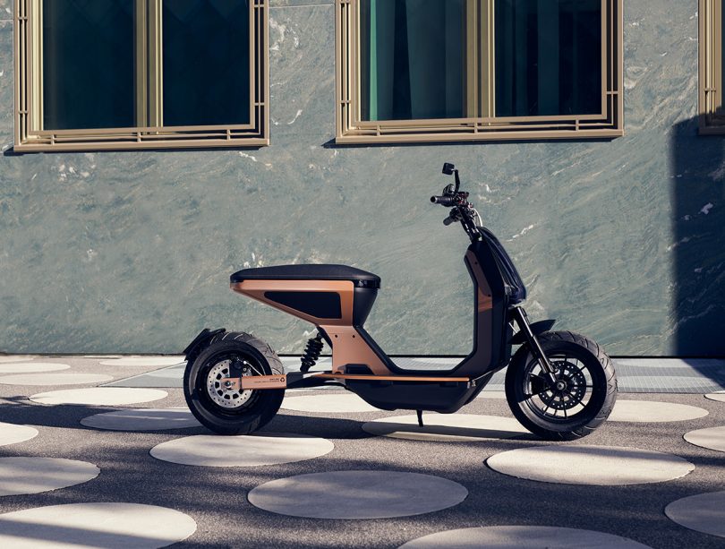 Side profile of rose bronze and black aluminum scooter parked against wall with window backdrop and ground with circular concrete.