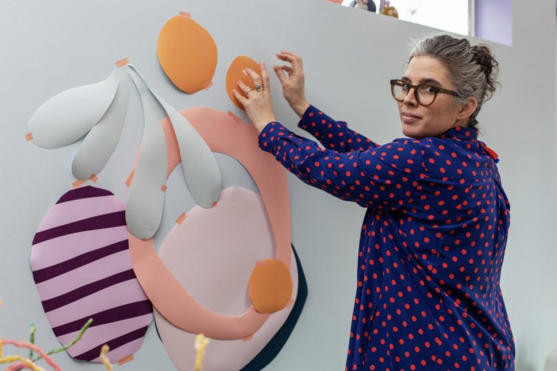 Artist Alex Proba looking toward camera over left shoulder while taping pieces of colored paper to wall, assembling a design motif for Samsung Bespoke refrigerator.