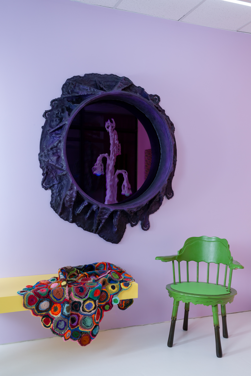 round purple-black mirror with amorphous frame on light purple wall, multicolored textile draped over yellow bench, and green wood armchair