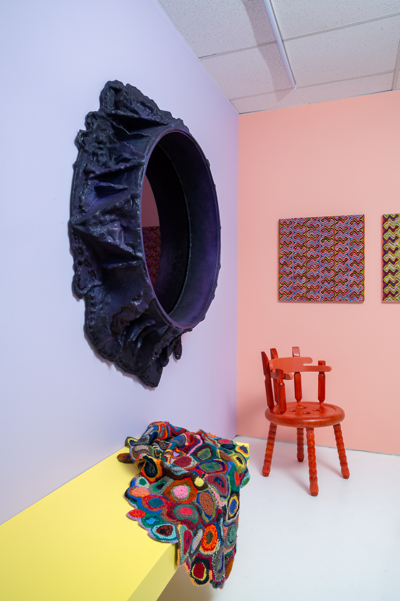 round purple-black mirror with amorphous frame on light purple wall, multicolored textile draped over yellow bench, and red wood armchair