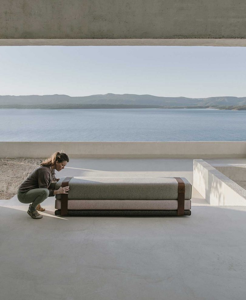 person crouched down looking at one end of an upholstered settee with a water landscape behind