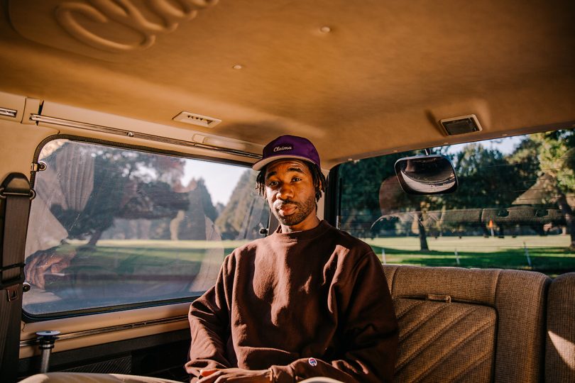 brown-skinned man sitting in the backseat of a car wearing a brown sweatshirt and hat