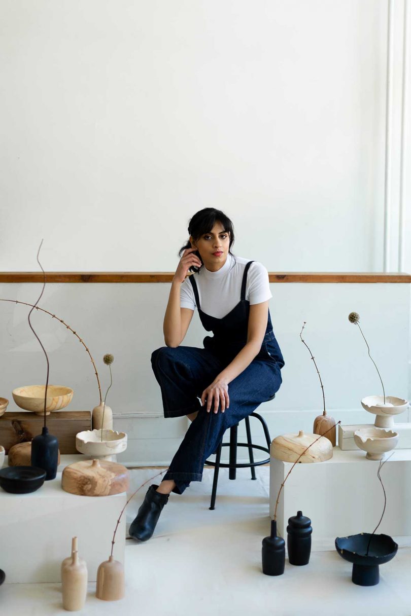 brown-skinned woman wearing dark jumpsuit over short sleeve white t-shirt sitting on a stool