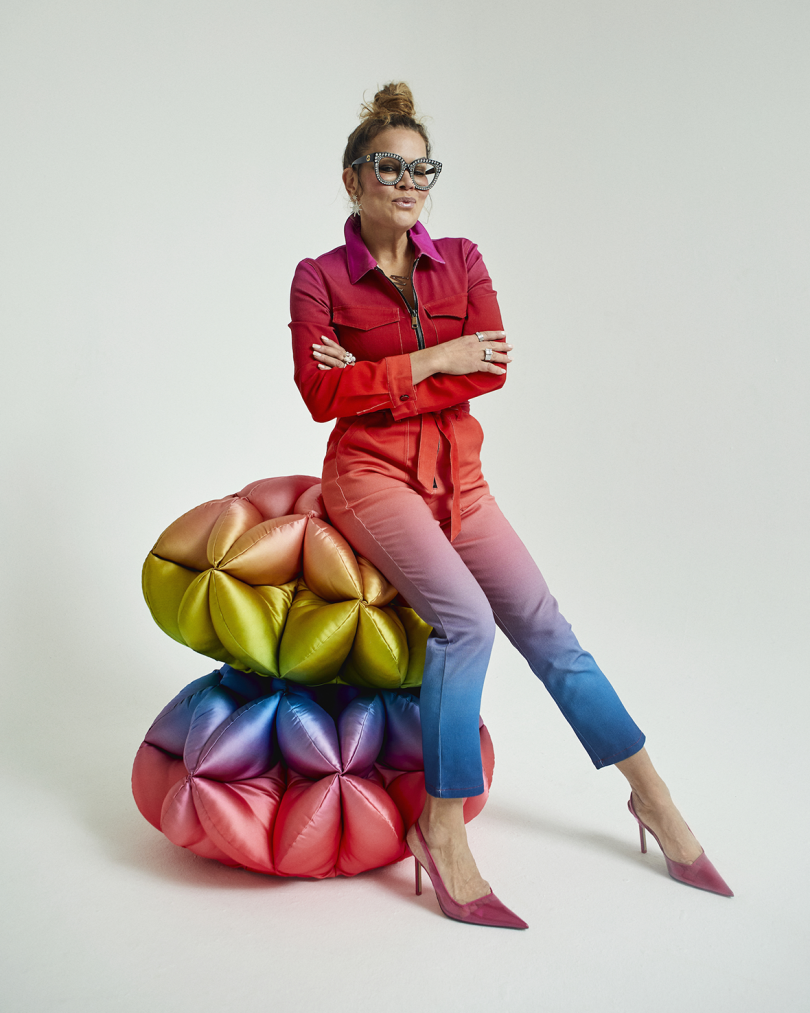 light-skinned woman wearing glasses and red and blue outfit leaning on two multicolored pouf