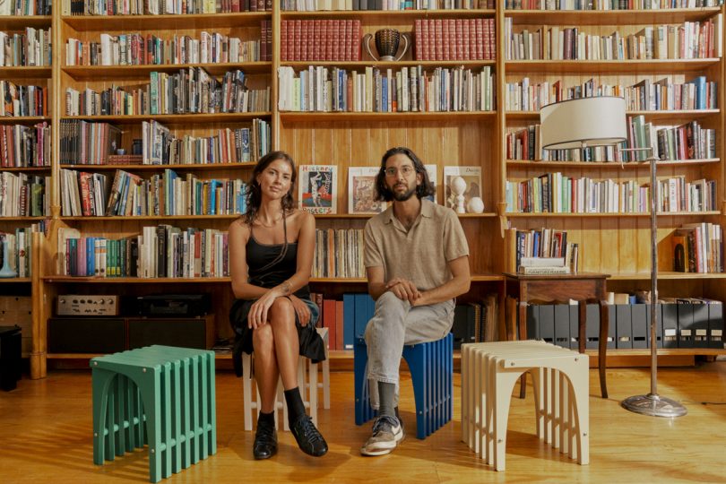 a light-skinned man and a light-skinned woman sitting on two stools in front of a wall of built-in bookshelves