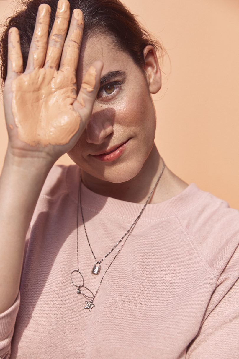 light-skinned woman wearing light pink shirt and necklaces holding up a hand with peach colored paint on the palm