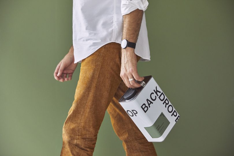 light-skinned man wearing white button down shirt and dark khaki pants carrying a gallon of Backdrop paint