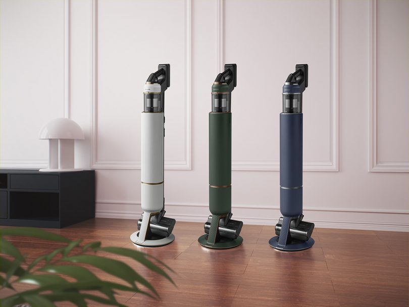 Three stick vacuums on stand in white, green and navy color options placed on wood floor with low bookshelf and white lamp near to the left and palm plant leaves in the foreground.