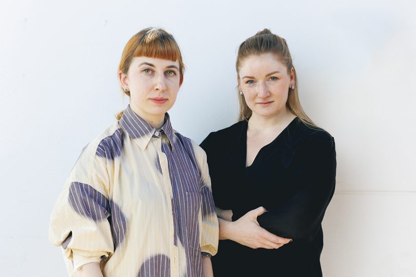 two light-skinned women with medium brown hair facing the camera with a white wall behind them