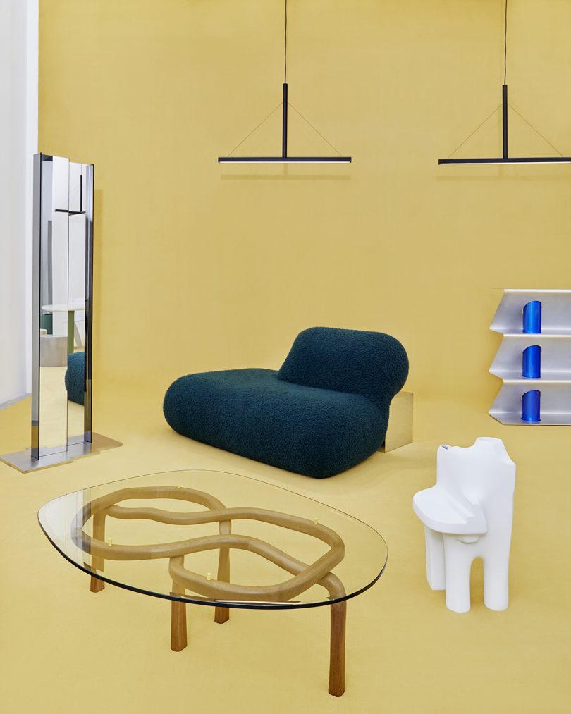 tall mirror, dark green chair, glass topped coffee table, shelving, and lighting on butter yellow background