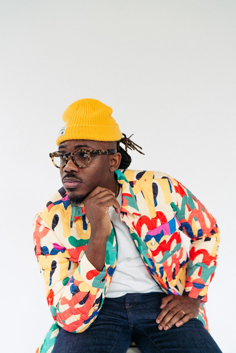 dark-skinned male wearing glasses, yellow hat, brightly colored patterned jacket looking off camera