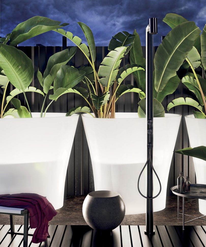slim black outdoor shower in front of three large potted palms