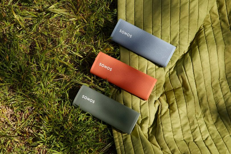Three Sonos Roam speakers in green, orange and blue on green picnic blanket and grass.