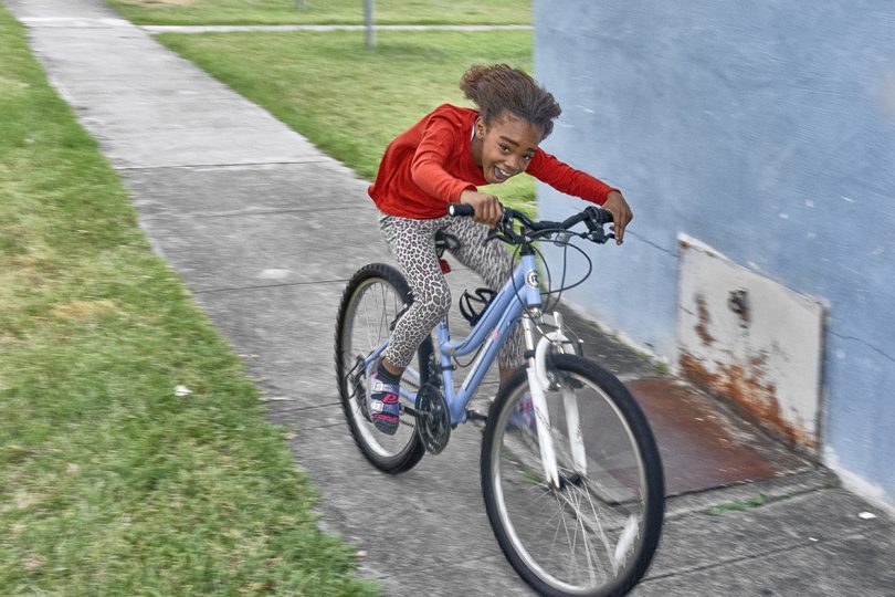 دختری با پوست تیره در حال دوچرخه سواری در پیاده رو و لبخند زدن